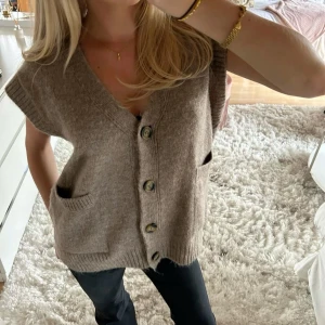 Beige stickad väst med knappar - Mysig beige stickad väst med v-ringning och stora knappar framtill. Västen har två fickor och ribbade kanter vid ärmhål och nederkant. Perfekt att slänga över en t-shirt eller skjorta för en avslappnad look.