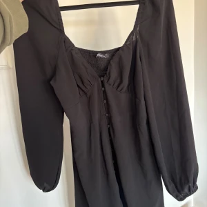 Svart klänning med knappar från Gina Tricot - Svart kort klänning från Gina Tricot med långa puffärmar, markerad midja och knappar framtill. Klänningen har en fyrkantig ringning och lätt rynkade detaljer vid bysten. Perfekt för dig som gillar en stilren och trendig look.