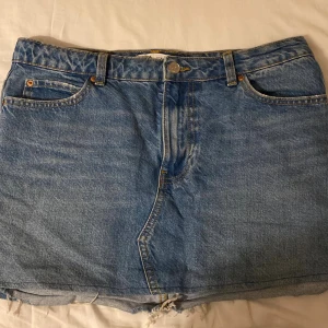 Blå jeanskjol med rå kant - Säljer en klassisk blå jeanskjol med fem fickor. Kjolen har bälteshällor, knapp och dragkedja framtill samt stora bakfickor. Materialet är slitstarkt denim med kontrastsömmar i gult. En fläck på insidan av fickan från ett läppglans och ett litet hål vid ett av bälteshällorna.