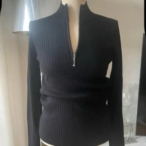 Svart ribbad tröja med half zip Soft Rebels ny med prislappen på  - Svart långärmad ribbad tröja från Soft Rebels med half zip framtill och hög krage. Slim passform som sitter snyggt mot kroppen. Perfekt för dig som gillar stilrena och enkla plagg med en modern touch. Materialet är mjukt och stretchigt, vilket gör den skön att bära.