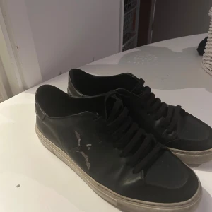 Svarta sneakers från Axel Arigato - Svarta sneakers från Axel Arigato i skinn med klassisk låg profil och vit sula. Skorna har snörning och en stilren design med få detaljer, förutom en fågelbrodyr på sidan. Perfekta för dig som gillar minimalistisk och clean stil. Kommer att tvättas innan ni köper. Kom privat efter köp för att se.