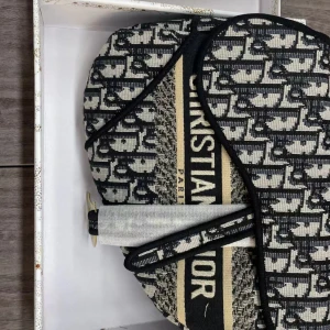 Dior Saddle Bag i canvas - Ikonisk Dior Saddle Bag i svart och beige canvas med tydligt logomönster och guldfärgade metalldetaljer. Väskan har en unik, böjd form och bred axelrem. Perfekt statement piece med broderad Christian Dior-text på framsidan.