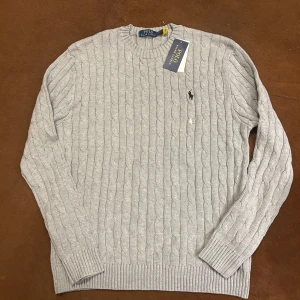 Grå kabelstickad tröja Polo Ralph Lauren - Kabelstickad grå tröja från Polo Ralph Lauren med klassisk rund halsringning och broderad logga på bröstet. Tröjan är långärmad och har ribbade muddar vid ärmslut och nederkant. Perfekt för lager-på-lager under kyliga dagar.