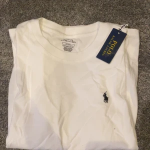 Vit t-shirt från Polo Ralph Lauren - Vit t-shirt från Polo Ralph Lauren med klassisk rund hals och svart broderad logga på bröstet. Modellen är kortärmad och har en clean, stilren look. Tillverkad i mjuk bomull för skön känsla hela dagen.