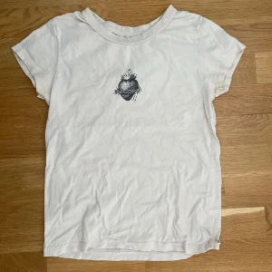 Vit t-shirt med hjärtmotiv från John Galt - Vit t-shirt från brandy Melville med ett svart tryck av ett hjärta med törnekrona och eld på bröstet. Klassisk rund halsringning och korta ärmar. Perfekt för en chill och stilren look.