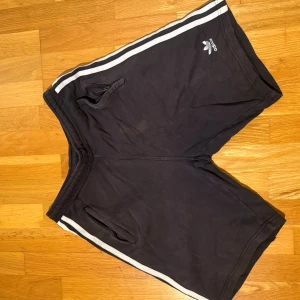 Svarta Adidas shorts med vita ränder - Svarta shorts från Adidas med klassiska vita ränder längs sidorna och Adidas-logga på ena benet. Modellen har två fickor med dragkedja och är tillverkad i mjuk bomull, perfekt för sport eller chill. Snygg och enkel design som funkar till det mesta.