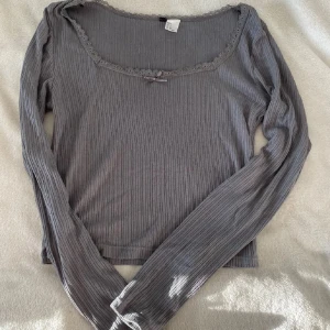 Grå ribbad långärmad topp med spets - Supersöt grå ribbad långärmad topp från H&M med spetskant runt halsen och en liten rosett framtill. Stretchigt material som sitter snyggt och har en feminin touch. Perfekt att styla med jeans eller kjol för en chill look.