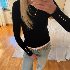 Svart långärmad topp med gulddetaljer - Snygg svart långärmad topp med rund hals och fyra guldknappar på ärmsluten. Modellen är slim och sitter tajt mot kroppen. Perfekt att styla med jeans eller kjol för en clean look. Materialet känns mjukt och stretchigt, troligen bomull eller polyester.