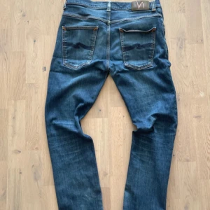 Blå jeans från Nudie Jeans, W34 L32 - Snygga blå jeans från Nudie Jeans med klassisk femficksdesign och orangea sömmar. Modellen har raka ben och normal passform, med coola detaljer på bakfickorna. Perfekt för dig som gillar stilrena och tidlösa jeans.