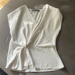 Vit omlottopp med knyt från Zara - Snygg vit omlottopp från Zara i storlek S. Toppen har v-ringning, knyt i sidan och är ärmlös. Materialet är mjukt och har en lätt glansig finish som ger en lyxig känsla. Perfekt för dig som gillar stilrena och trendiga plagg.