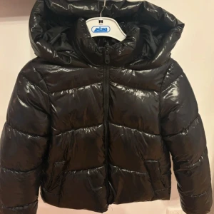 Svart pufferjacka Vero Moda XS - Säljer en svart, glansig pufferjacka från Vero Moda i storlek XS. Jackan har en stor huva, dragkedja framtill och två sidofickor. Den är quiltad och har långärmade ärmar med elastiska muddar. Perfekt för kalla dagar och har en loose passform.