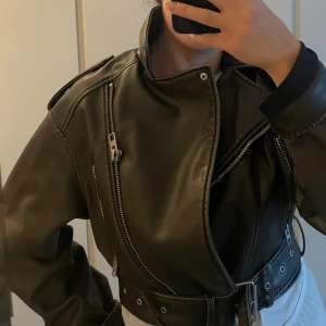 Mörkgrön  skinnjacka med bälte och dragkedjor - Säljer en mörkgrön  skinnjacka i bikerstil med coola detaljer som bred krage, flera silvriga dragkedjor och ett brett bälte i midjan. Jackan har en oversized passform och långa ärmar, perfekt för dig som vill ha en edgy look.