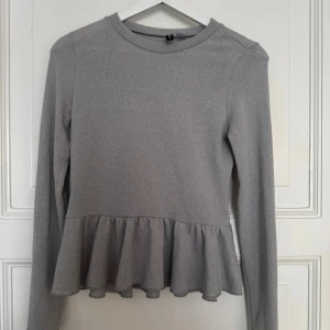 Glittrig silver topp med volang från H&M - Supersnygg silverfärgad topp från H&M Divided med glittrigt tyg och volang nertill. Långärmad och rund halsringning, perfekt för dig som vill sticka ut lite extra. Materialet är mjukt och stretchigt för en bekväm passform.