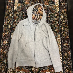 Grå hoodie från Burberry med rutig huva - Säljer en grå hoodie från Burberry i bomull med dragkedja och klassisk rutig insida i huvan. Tröjan har broderad logga på bröstet, snörning i huvan och två fickor framtill. Perfekt för dig som vill ha en stilren men ändå lyxig look. Pris kan diskuteras vid snabb affär