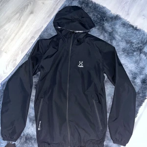 Svart vindjacka från Haglöfs  - Svart vindjacka från Haglöfs med huva och hel dragkedja framtill. Jackan har två sidofickor med dragkedja och meshfoder på insidan. Perfekt för blåsiga dagar och outdoor-aktiviteter. Klassisk och stilren design med diskret logga på bröstet.