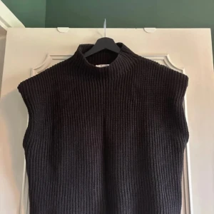 Svart stickad väst från NA-KD - Säljer en svart, ribbstickad väst från NA-KD med hög krage. Västen har en loose passform och är perfekt att slänga över en skjorta eller t-shirt för en chill och trendig look. Materialet är mjukt och stickat, vilket gör den både snygg och bekväm.