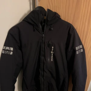 Svart Sail Racing vindjacka med huva - Svart vindjacka från Sail Racing med huva och dragkedja framtill. Jackan har logga på bröstet och båda ärmarna, samt flera praktiska fickor. Materialet är syntet och jackan har quiltad insida för extra komfort. Perfekt för blåsiga dagar. Storlek S. Pris kan diskuterar 