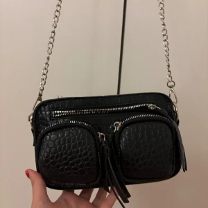 Svart axelväska med kedja och crocomönster - Snygg svart axelväska med crocomönstrad yta och silverfärgad kedjerem. Väskan har två framfickor med dragkedja och tofsdetaljer samt en extra dragkedja upptill. Perfekt storlek för det viktigaste och ger en cool vibe till outfiten.