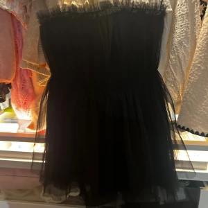 Svart axelbandslös klänning/topp H&M - Supersnygg svart axelbandslös kortklänning/topp från H&M med volangdetalj upptill och lager av tunn tyll som ger extra volym. Perfekt för dig som vill sticka ut med en klassisk men trendig look. Klänningen/toppen har en markerad midja och är helsvart.