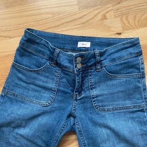 Blå bootcut jeans från Nelly - Snygga blå jeans från Nelly med bootcut-modell och hög midja. Jeansen har dubbla knappar framtill, stora framfickor och bakfickor med lock och knapp. Tillverkade i klassiskt denimtyg med en lätt tvättad look.