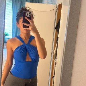 Blå omlott topp från Zara - Säljer en blå omlott topp från Zara i storlek S. Toppen har en snygg twistad detalj framtill, är ärmlös och har en tight passform. Materialet är stretchigt och känns mjukt mot huden. Använd ett fåtal gånger och är lite nopprig baktill i nacken.