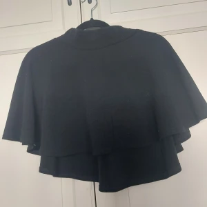 Svart bolerotopp med volang - ⚠️ Skriv till mig innan köp!⚠️Säljer en svart minikjol med dubbla lager och volangig design. Toppen har en högre halsringning och är tillverkad i ett mjukt, stickat material. Perfekt för att slänga över en klänning eller topp för en cool lager-på-lager-look.