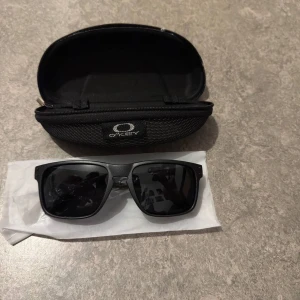 Svarta Oakley Holbrook solglasögon - Snygga svarta Oakley Holbrook solglasögon med klassisk fyrkantig form och mörka linser. Kommer med original hårt fodral med dragkedja. Perfekta för dig som vill ha en stilren och sportig look.