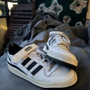 Adidas Forum Low vita och svarta sneakers - Säljer ett par klassiska Adidas Forum Low sneakers i vitt skinn med svarta detaljer och tre ränder på sidorna. Skorna har snörning, kardborreband och svart yttersula. Perfekta för dig som gillar retrostil och streetwear.