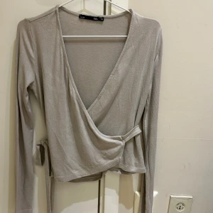 Beige omlottopp från 157 XS - Säljer en beige omlottopp från 157 i storlek XS. Toppen har långa ärmar, v-ringning och knyt i sidan för en snygg passform. Perfekt att styla med jeans eller kjol för en trendig look.
