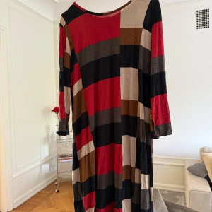 Randig långklänning med dragkedja - Långklänning med breda blockrandiga mönster i rött, svart, beige, brunt och grått. Klänningen har trekvartsärm och en synlig dragkedja bak. Tillverkad i ett mjukt, lätt material som faller snyggt. Perfekt för dig som gillar färg och grafiska mönster.