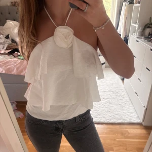 Vit volangig halterneck topp - Supersöt vit halterneck topp med dubbla volanger och en stor tygblomma framtill vid halsen. Toppens material är lätt och luftigt, perfekt för varma dagar. Den är ärmlös och har öppen rygg, vilket ger en fräsch och trendig look.