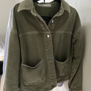 Olivgrön jeansjacka från Zara - Snygg olivgrön jeansjacka från Zara med klassisk krage, stora framfickor och knäppning framtill. Jackan har en avslappnad passform och är tillverkad i kraftigt bomullstyg. Perfekt för dig som gillar en stilren och enkel look.