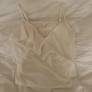 Vit omlott linnetopp med volang - Säljer en vit linnetopp från H&M i storlek M. Toppen har smala axelband, omlott-design och en snygg volang som går diagonalt över framsidan. Perfekt för sommarens varma dagar och har en feminin och fräsch look.