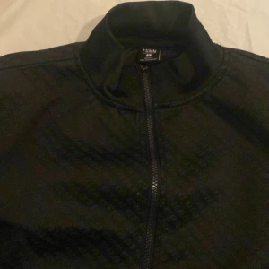 Svart chaotic hoodie - Säljer en svart hoodie  fmed diskret mönster och hel dragkedja framtill. Hoodien  har en klassisk bomberkrage och är tillverkad i ett mjukt syntetmaterial. Perfekt för dig som vill ha en stilren och enkel hoodie.