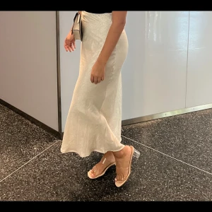 Vit långkjol i satin - Säljer en elegant vit långkjol i skimrande satin med lätt utsvängd passform. Kjolen har en slät midja och följsam siluett som ger en stilren look. Perfekt för dig som gillar en clean och chic stil. Passar S/M