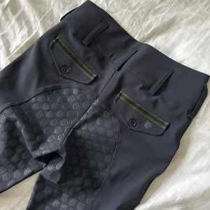 Svarta ridbyxor med mönstrade detaljer från Maya Delorez - Snygga svarta ridbyxor med grått mönster på insidan av benen för extra grepp. Byxorna har gröna detaljer vid fickorna och knäppning fram. Perfekta för dig som vill ha stil och funktion i stallet.