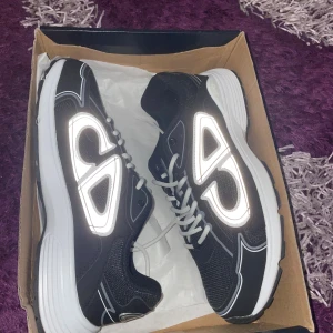 Svarta Dior B30 sneakers med reflexdetaljer - Säljer ett par svarta Dior B30 sneakers med feta reflexdetaljer på sidorna och coola CD-loggor. Skorna har vit och svart sula, mesh och syntetmaterial samt vita snören. Perfekta för dig som vill sticka ut med en lyxig och sportig vibe.