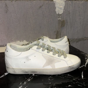 Golden Goose vita sneakers med stjärna - Golden Goose sneakers i vitt skinn med grå mockadetaljer och ikonisk stjärna på sidan. Skorna har grå snörning, rund tå och platt sula. Snyggt slitna detaljer på sulan och hälen ger en cool, avslappnad vibe. Perfekta för dig som gillar streetstyle och unika detaljer.