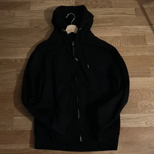 Svart hoodie från Pull&Bear med dragkedja - Svart hoodie från Pull&Bear i storlek M. Tröjan har huva med snörning, hel dragkedja framtill och ribbade muddar vid ärmslut och nederkant. Perfekt för chill dagar och enkel att matcha med jeans eller joggers.