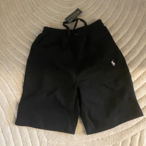 Svarta shorts från Polo Ralph Lauren - Svarta shorts från Polo Ralph Lauren med klassisk vit logga broderad på ena benet. De har elastisk midja med snörning och är tillverkade i mjuk bomull, perfekta för en avslappnad stil.