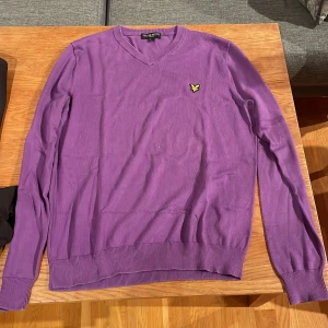 Lila stickad tröja från Lyle & Scott - Säljer en lila stickad tröja från Lyle & Scott med klassisk v-ringning och gul broderad logga på bröstet. Tröjan har långa ärmar och ribbade muddar vid ärmslut och nederkant. Perfekt för dig som vill ha en färgstark och stilren look.