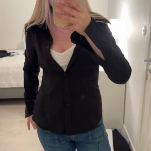 Svart figursydd skjorta med knappar - Svart skjorta med klassisk krage och knäppning framtill. Figursydd modell som sitter snyggt på kroppen och har långa ärmar. Perfekt att styla med jeans eller kjol för en clean look.