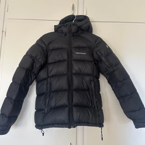 Svart dunjacka Peak Performance - Svart dunjacka från Peak Performance med huva och dragkedja framtill. Perfekt för kalla dagar med sin fluffiga och varma design. Justerbar nedtill med snörning. Sparsamt använd. 