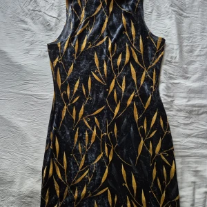 Mönstrad klänning, I svart och guld, från Savann - Snygg ärmlös svart klänning från Savann med guldigt bladliknande mönster. Klänningen är figurnära och tillverkad i mjuk polyester med en lätt glansig finish. 