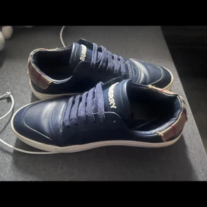Mörkblå Burberry sneakers i skinn - Snygga mörkblå sneakers från Burberry i skinn med klassiskt rutigt tygdetalj vid hälen. Skorna har vita sulor, rund tå och snörning framtill. Perfekta för dig som vill ha en stilren och lyxig look med en touch av ikoniskt mönster. Nypris 3500