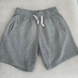 Grå mjuka shorts med snörning - Säljer ett par grå shorts i mjukt material med elastisk midja och vit snörning framtill. Shortsen har två sidofickor och en avslappnad passform, perfekta för chill dagar eller träning.