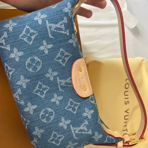 Louis Vuitton denim axelväska monogram - Säljer en snygg axelväska från Louis Vuitton i blått denimtyg med ikoniskt monogrammönster. Väskan har ljusa skinn-detaljer, röd kant och guldfärgade metalldetaljer. Stängs med dragkedja och har en stilren, kompakt form som är lätt att bära.