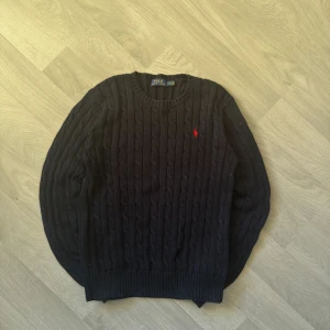 Mörkblå kabelstickad tröja Polo Ralph Lauren - 100% äkta- marinblå tröja från Polo Ralph Lauren -  klassisk röd broderad logga på bröstet - 100% bomull - rund halsringning - Perfekt för en stilren och tidlös look.