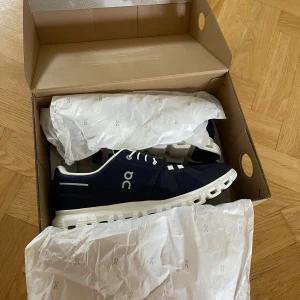 Marinblå On Cloud sneakers - Säljer ett par marinblå On Cloud sneakers med vit och svart sula. Skorna har mesh-ovandel, vita snören och On-logga på sidan. Snygg och sportig design med unik sula för extra komfort. Perfekta för dig som gillar stilrena och tekniska sneakers. Skorna är helt nya köpte fel storlek 