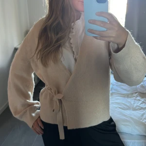 Beige omlottkofta med knytband - Superfin beige omlottkofta med knytband i midjan och långa ärmar. Koftan har en mjuk och fluffig känsla och är perfekt för lager-på-lager. Passar snyggt till både kjol och jeans.